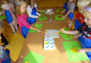 Dzieci stoją przy stoliku i malują liście żółtą farbą